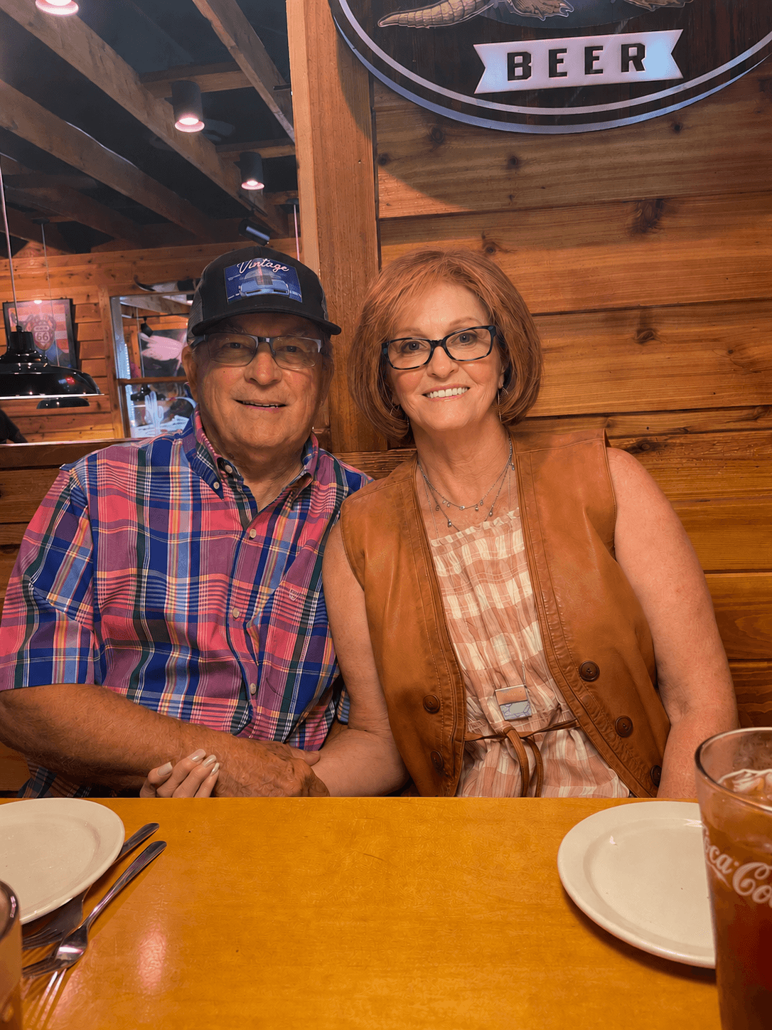 A man and woman sit at a wooden table in a restaurant, smiling at the camera. The man wears glasses and a plaid shirt, the woman has glasses and a sleeveless vest.