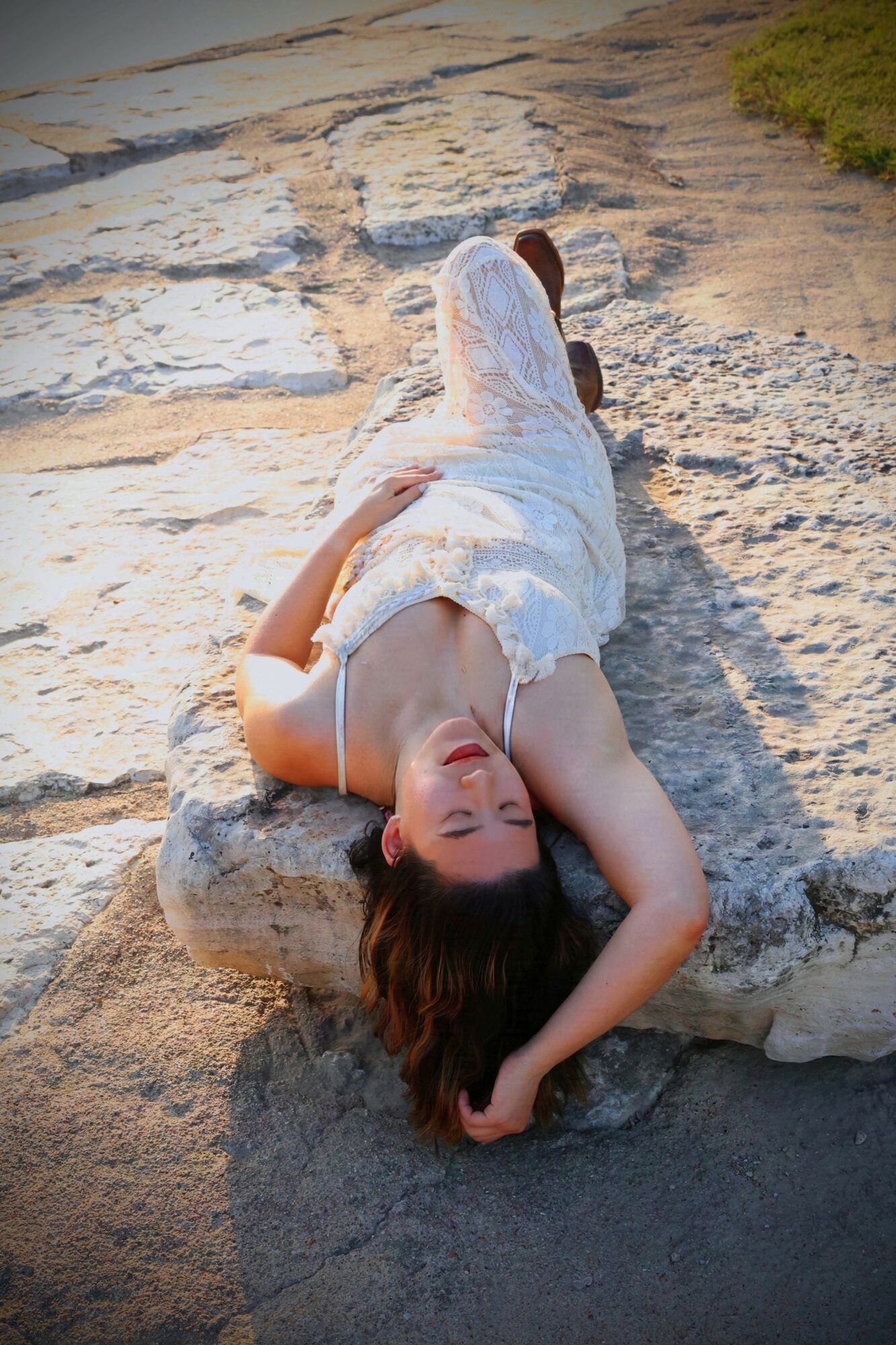 Woman lying on a large rock outdoors, with her head resting on her arm, wearing a white dress, sunlit scene.