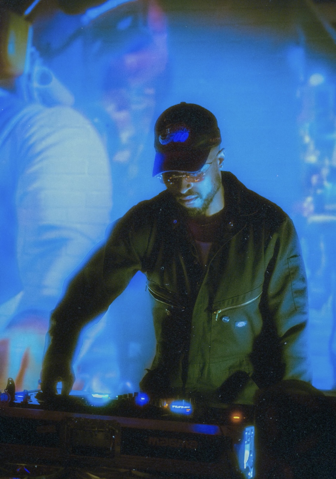 Person wearing a cap and jacket, standing behind a DJ console, with blue lighting and reflections in the background.