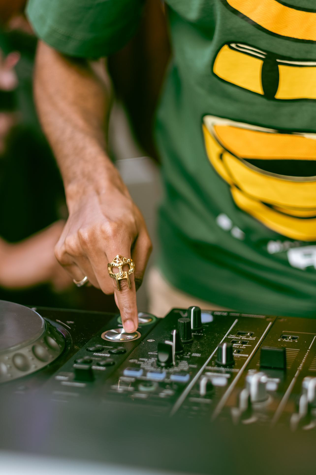 Person's hand adjusting DJ equipment, wearing a green shirt with yellow and black design, ring on finger.