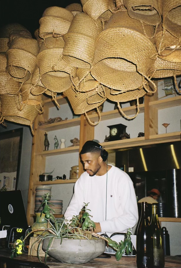 Chef wearing headphones and white uniform preparing food in a modern kitchen with hanging woven baskets overhead.