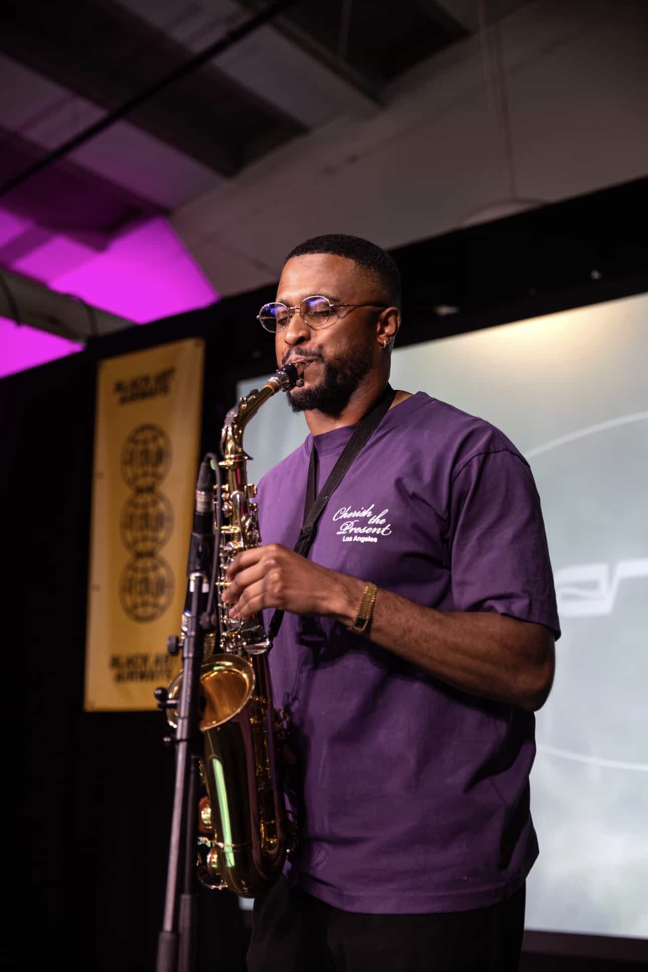 Man playing saxophone on stage, wearing glasses and a purple shirt, with a yellow banner in the background.
