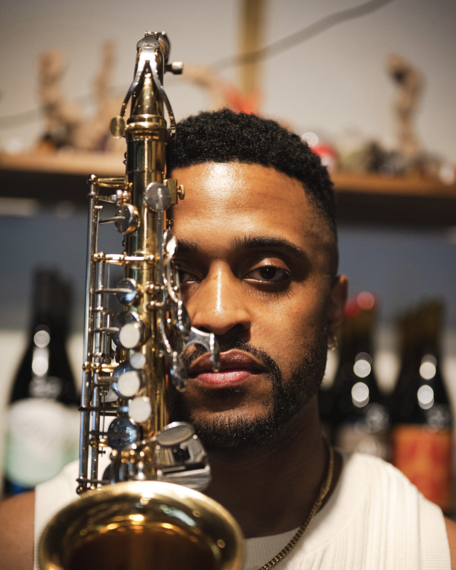 A man with short curly hair and a beard holds a gold-colored saxophone in front of his face, with a blurred background.