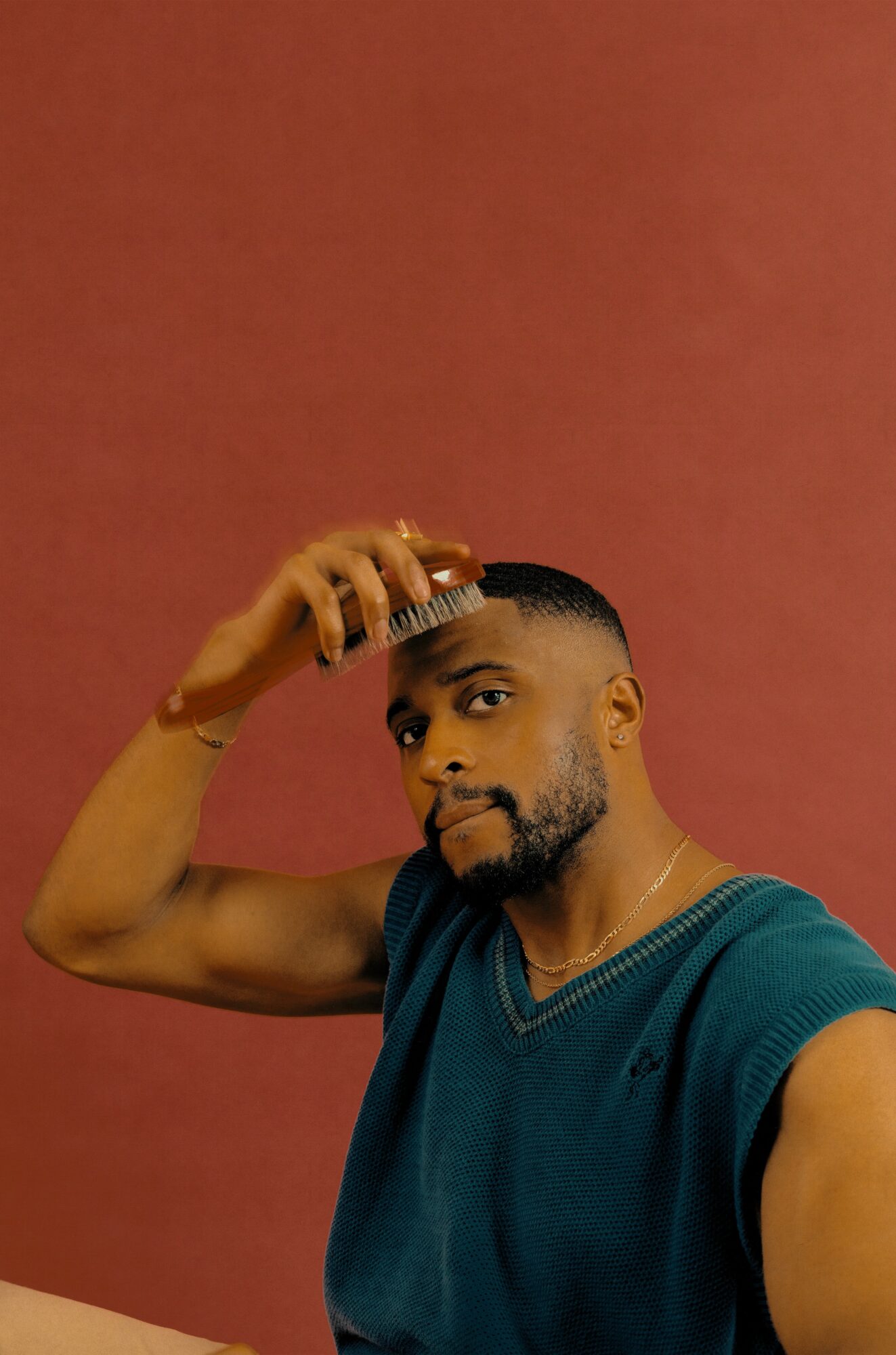 Man with short hair and beard brushes hair with a comb, wearing a blue sleeveless shirt, against a reddish background.