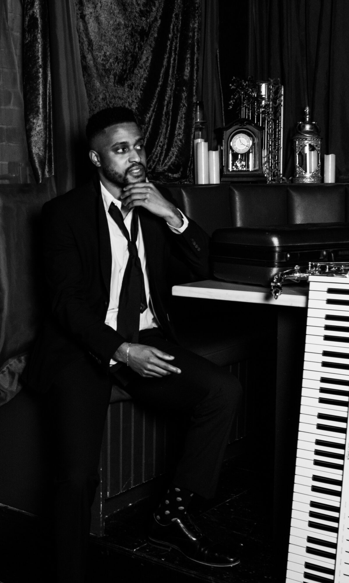 Man in suit sitting at piano with hand on chin, in a room with curtains and decorative items.
