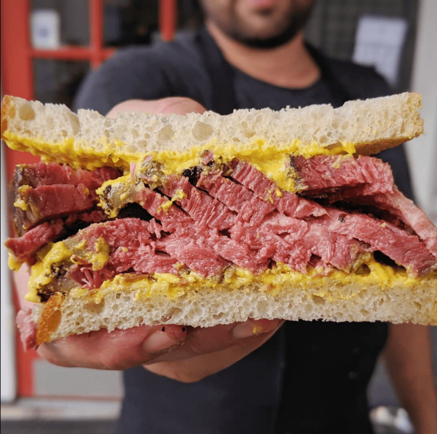 Person holding a large sandwich with bread, cooked meat, and egg, in front of a blurred background.