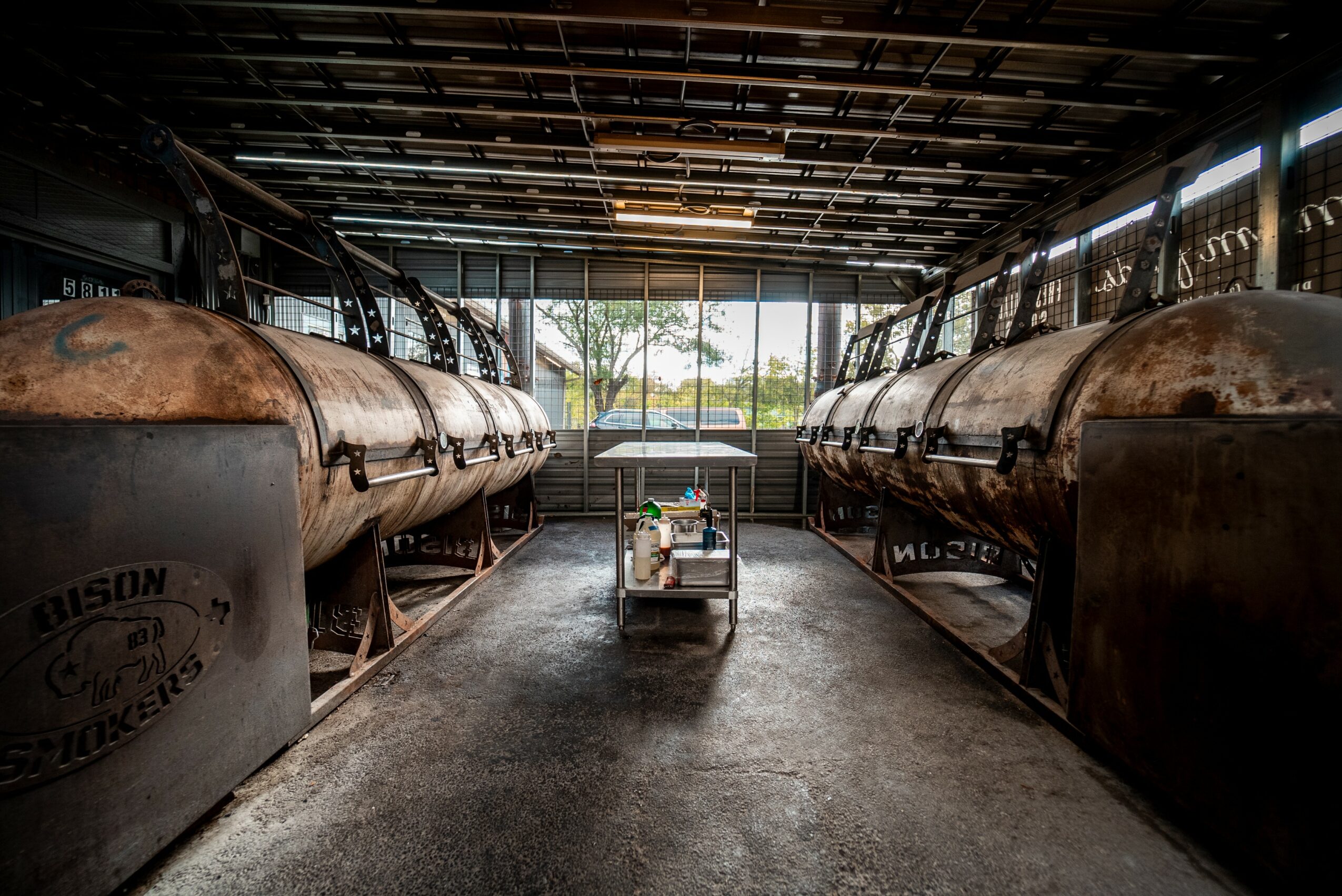 Industrial space with large rusted tanks on both sides and a cart in the center, with a window showing trees outside.