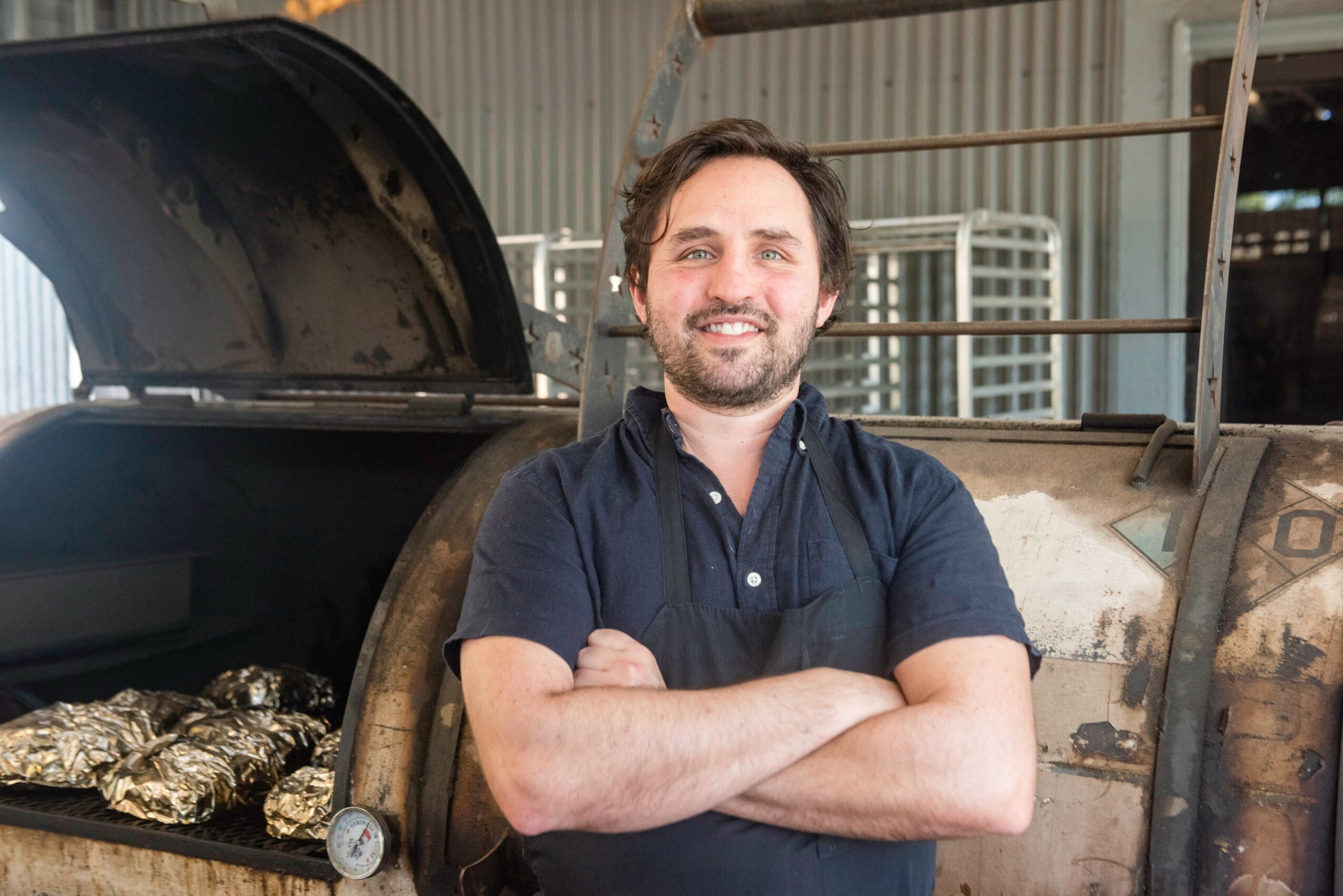 Man with dark hair and beard smiling with arms crossed, standing outdoors near a large industrial oven or smoker.