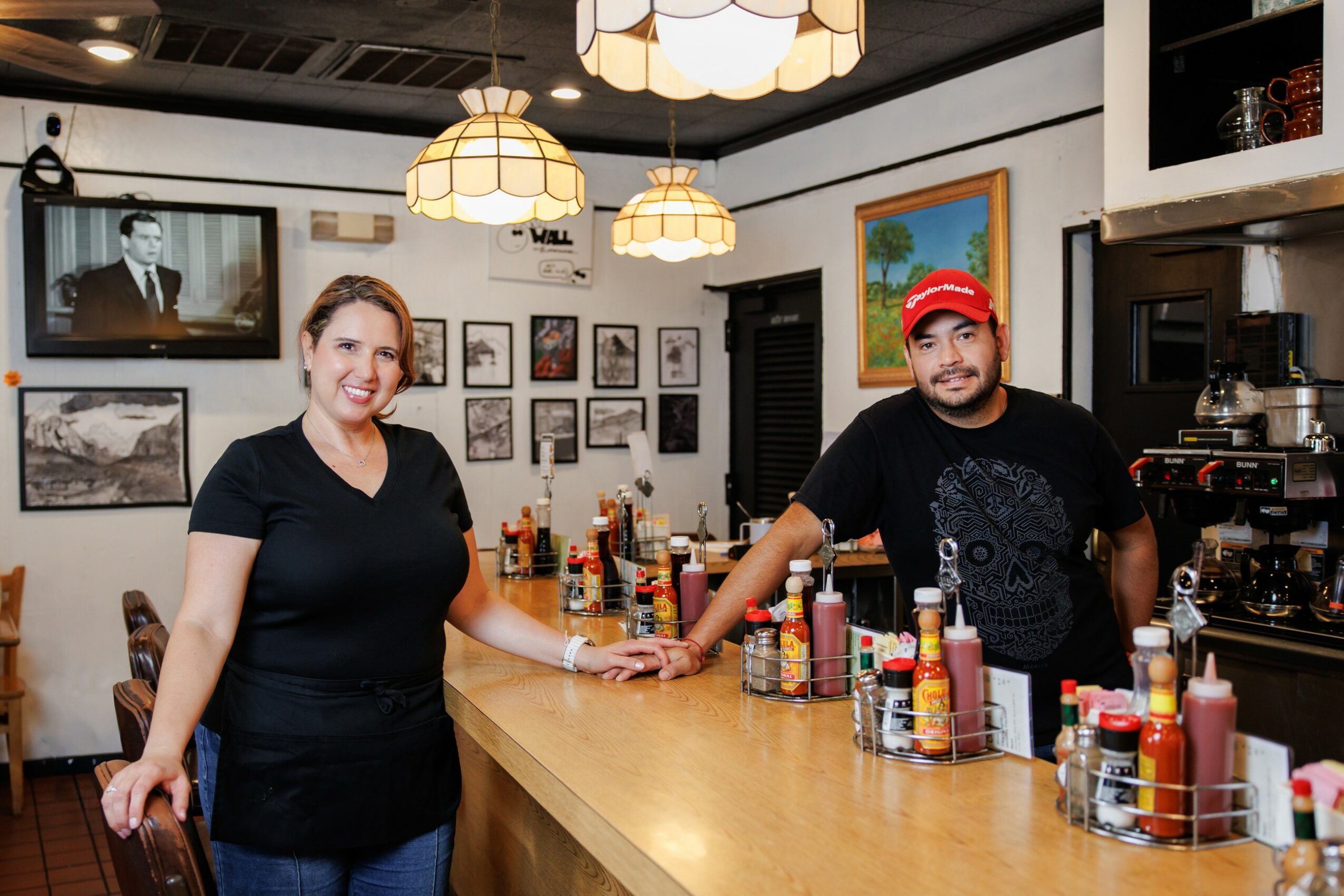 Two people behind a bar counter with bottles, smiling, in a cozy room with framed pictures and hanging lights.