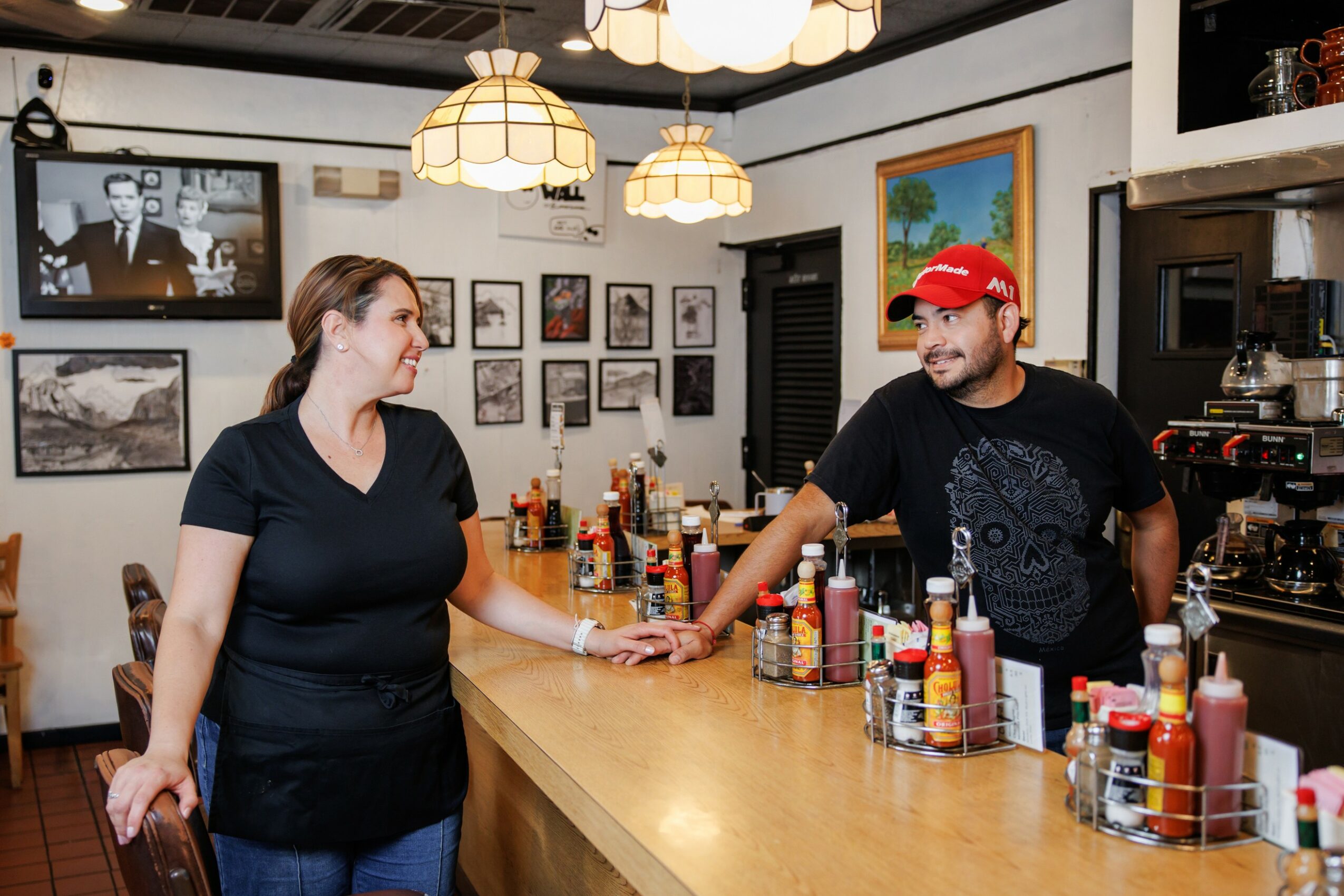 A woman and a man are talking at a bar counter with bottles and condiments, in a cozy restaurant setting.