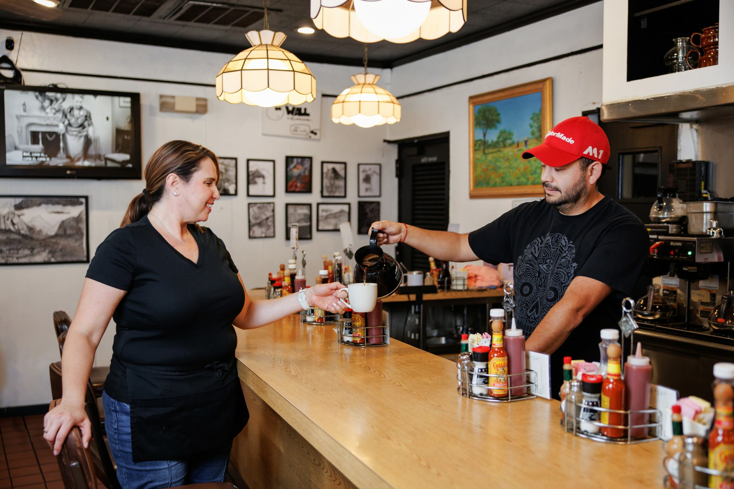 Woman at bar receiving drink from bartender in a cozy restaurant with framed pictures and paintings, warm lighting.