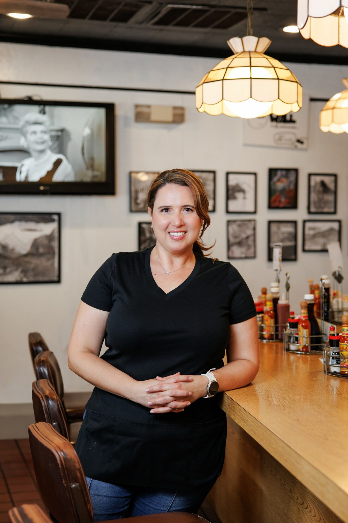 Woman standing behind a bar counter in a restaurant, smiling, with framed pictures on the wall behind her.