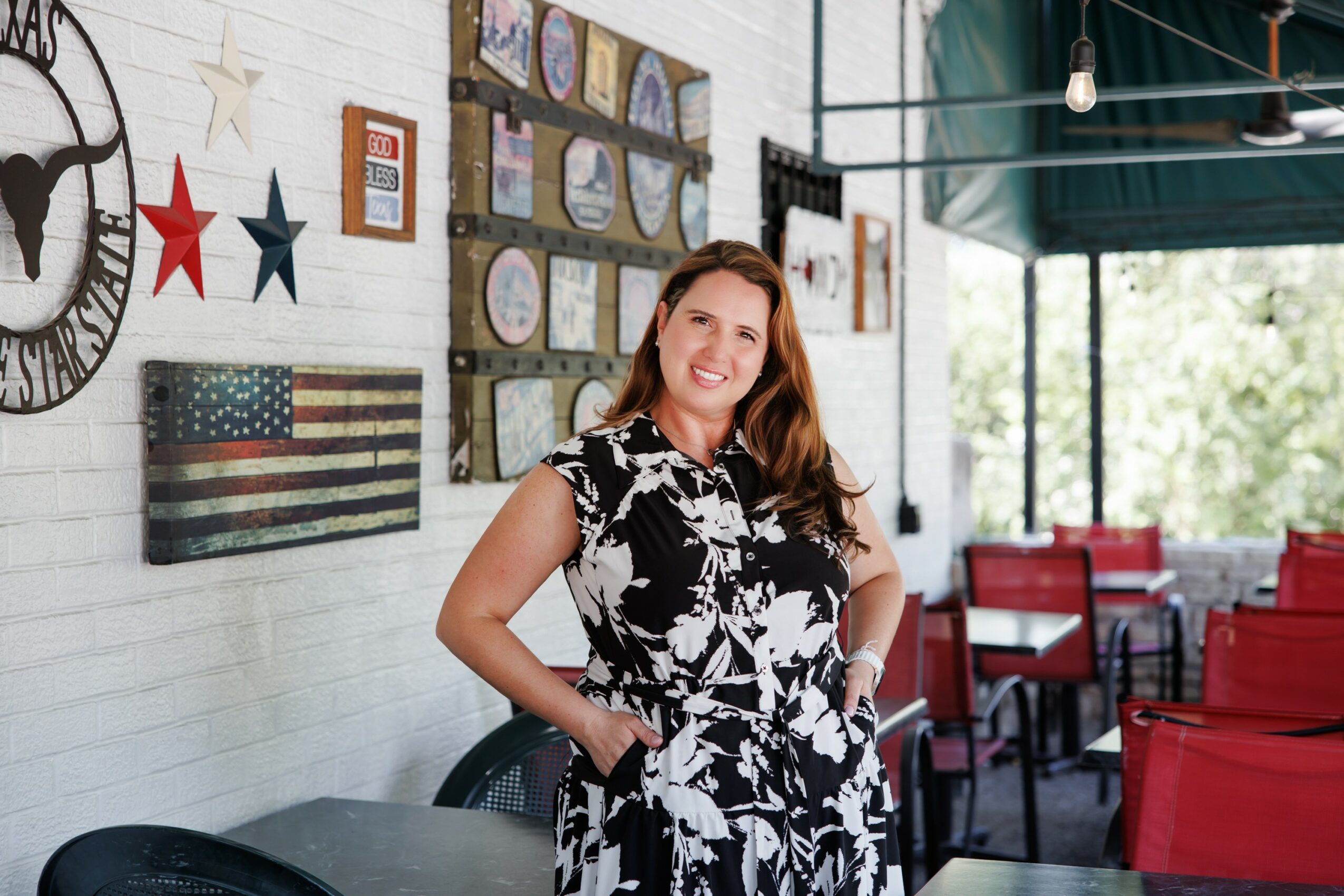 Smiling woman with long hair in a black and white floral dress, standing indoors near a white brick wall with patriotic decorations.