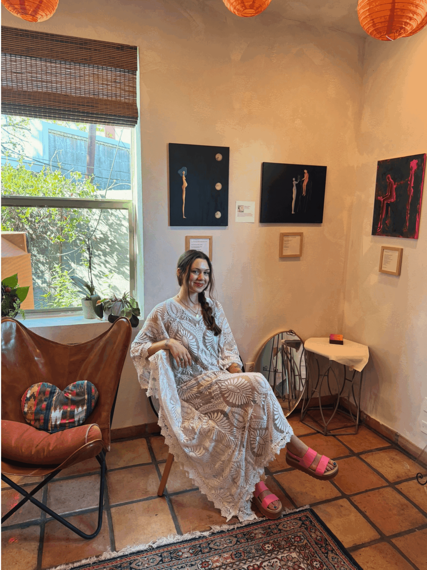 Woman sitting on a chair in an art gallery, surrounded by paintings and a window with sunlight, wearing a patterned dress and pink shoes.