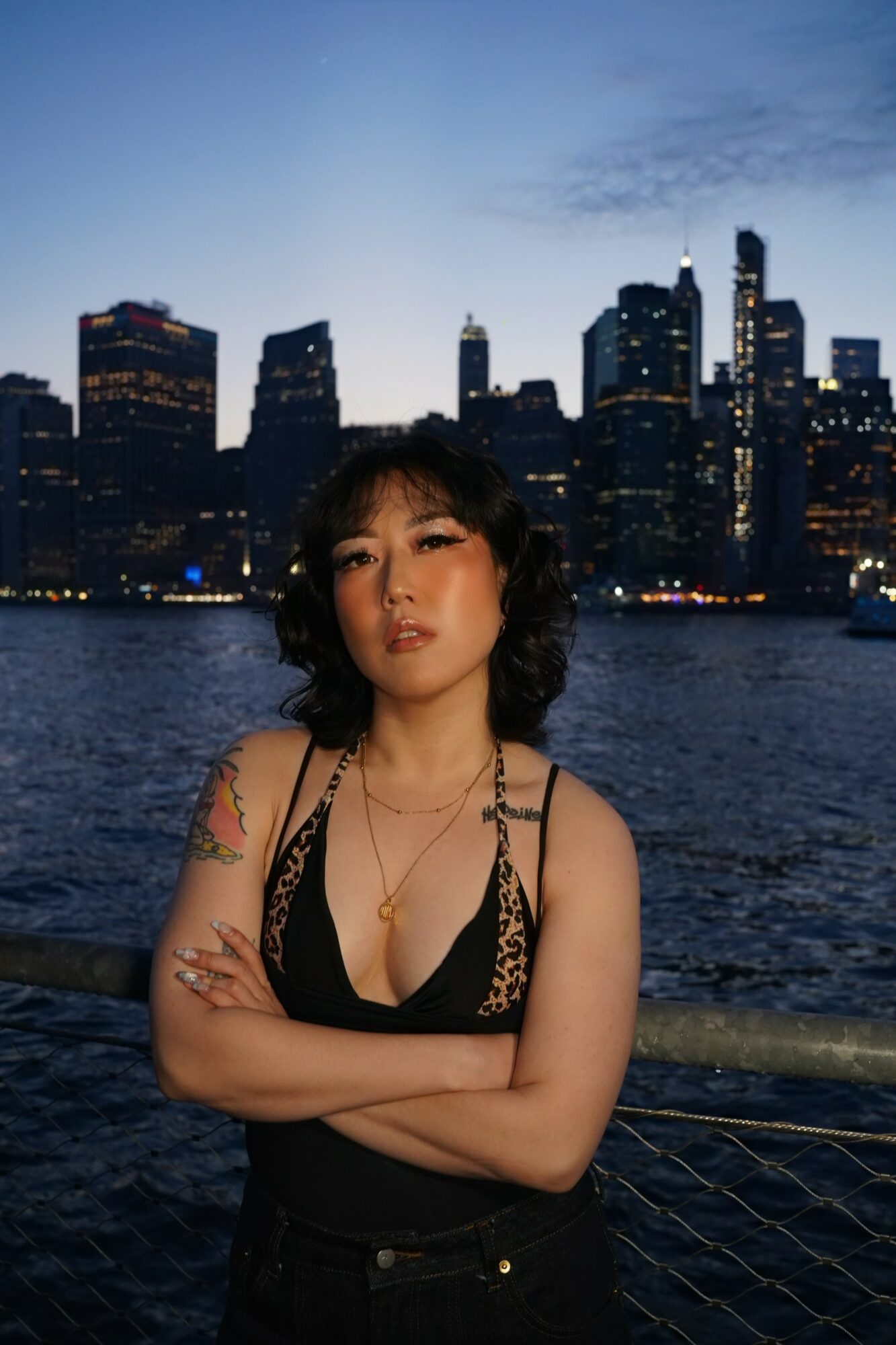 Young woman with dark hair and tattoos standing by water with city skyline at dusk.