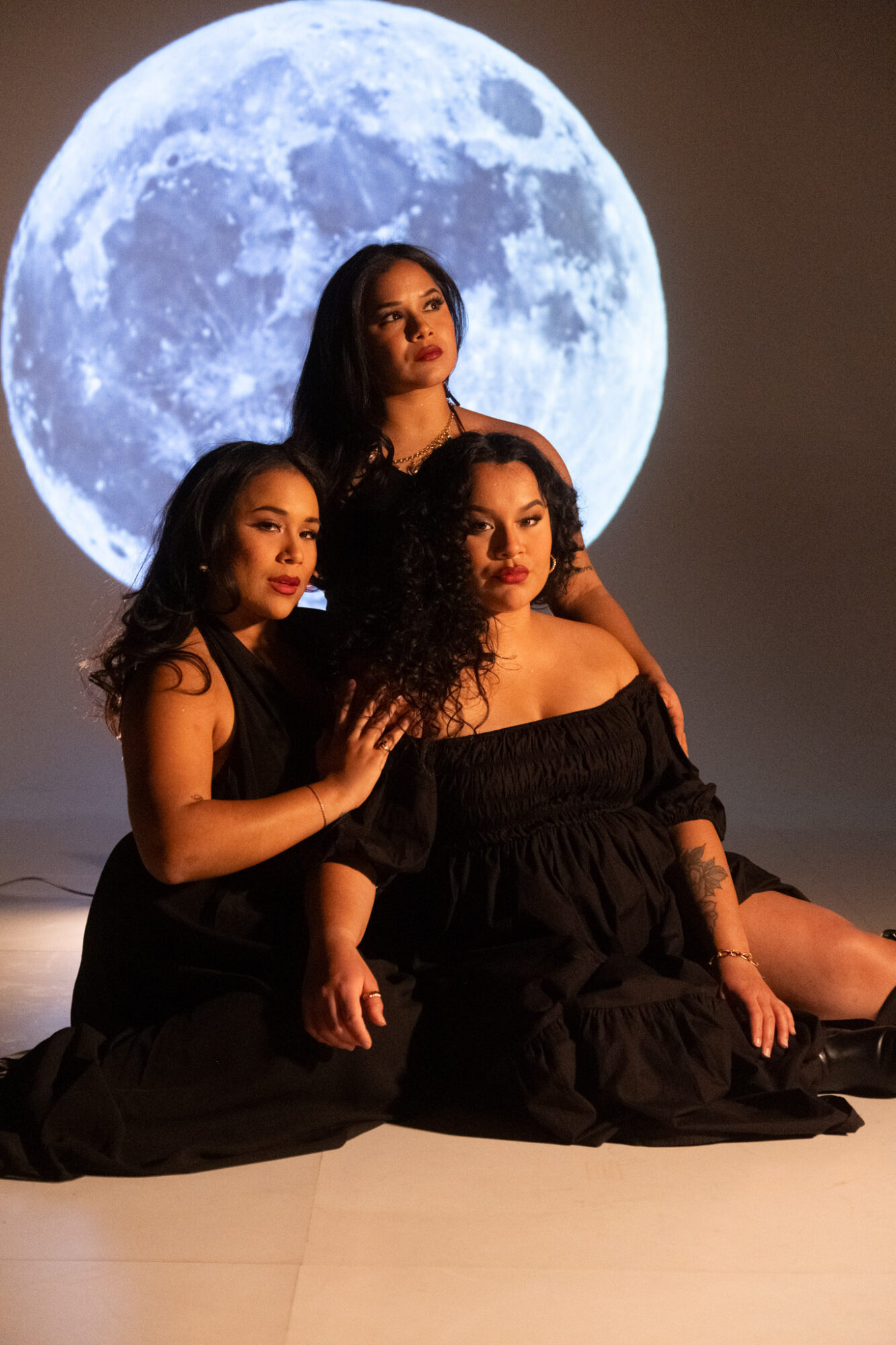 Three women in black dresses sit on the floor with a large moon backdrop behind them.