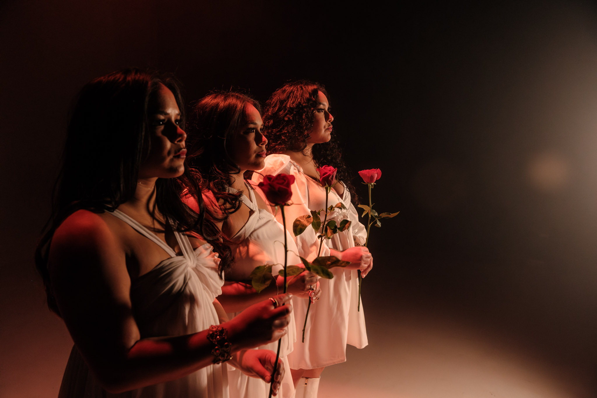 Four women in white dresses holding pink roses, standing in a dark setting with a light source on the right.