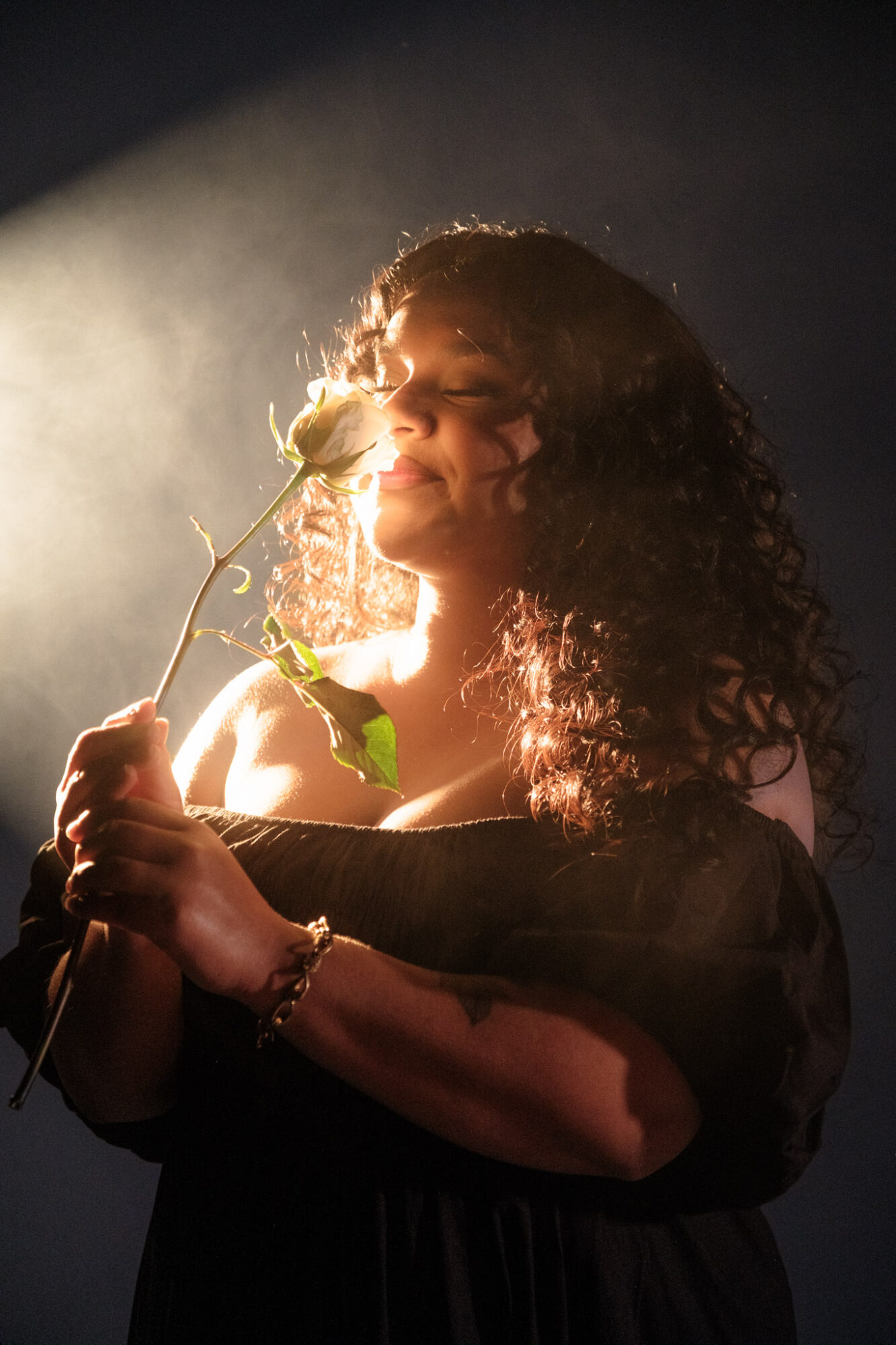 Woman with curly hair holding a flower, smiling with closed eyes, backlit by soft light.