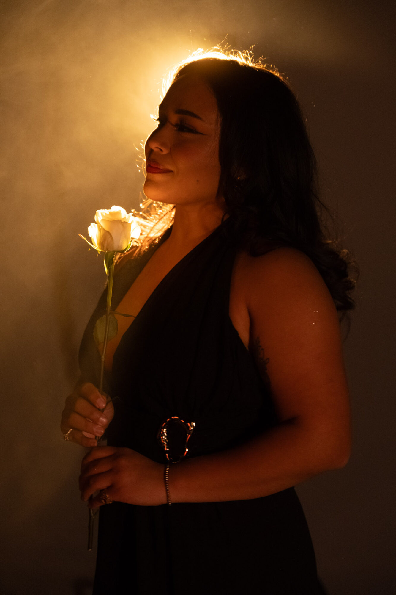 Woman holding a white rose, facing right, with backlit hair and a dark dress, in warm lighting.