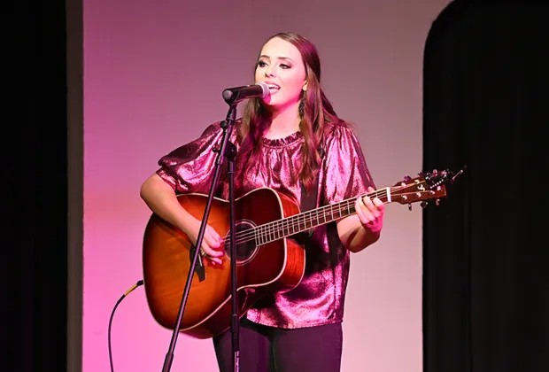 Young woman singing and playing guitar on stage with pink background and black curtain, wearing a shiny pink top.