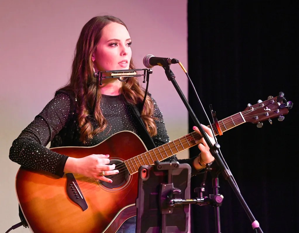 Young woman with long hair playing an acoustic guitar and singing into a microphone on stage.