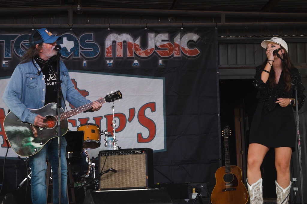 Two women perform on stage, one playing guitar and the other singing into a microphone, with a banner and musical equipment behind them.