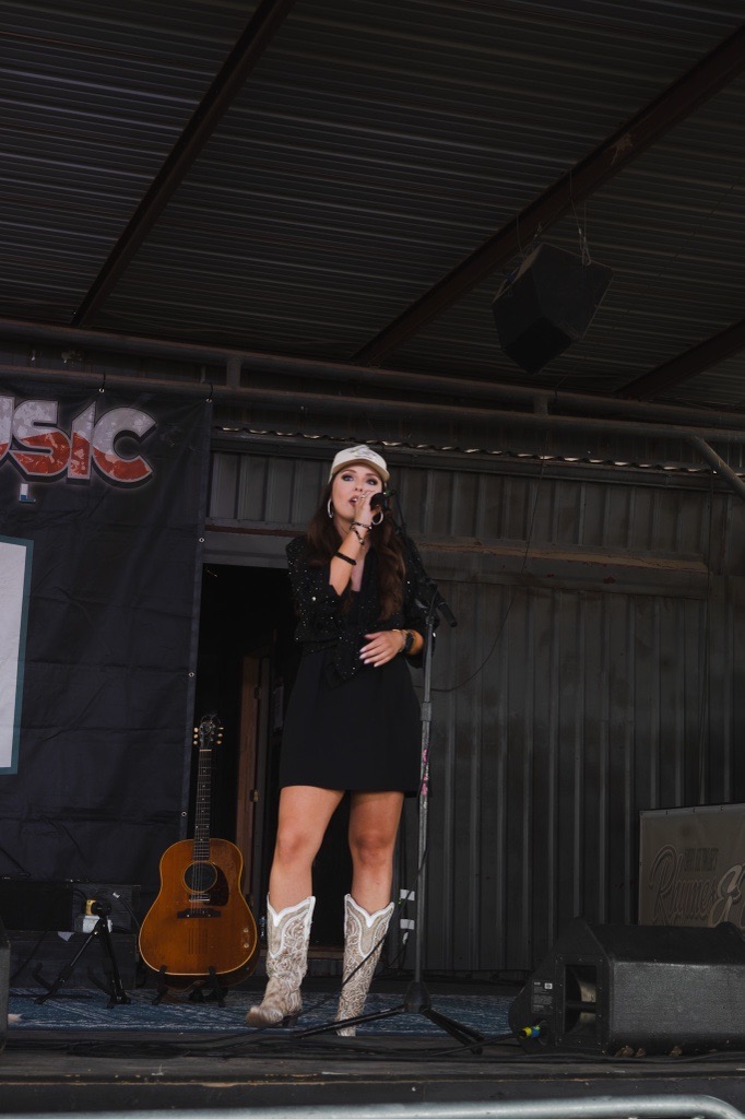 A woman in a black dress and white boots singing on stage with a guitar behind her.