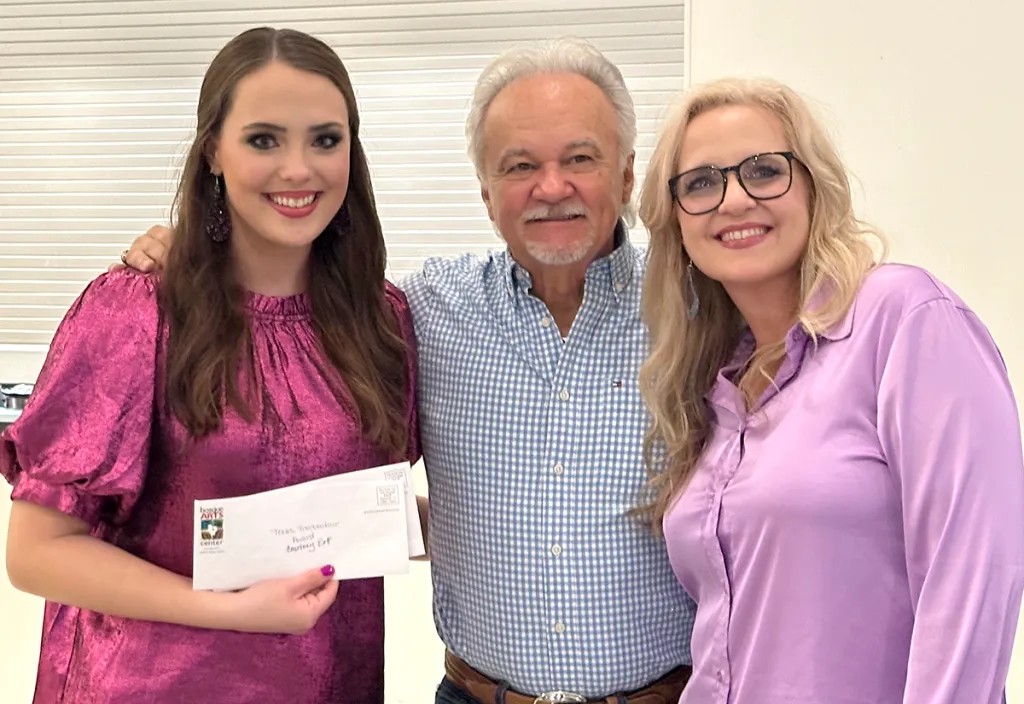 Three people smiling, woman on left holding a check, standing indoors with blinds behind them.
