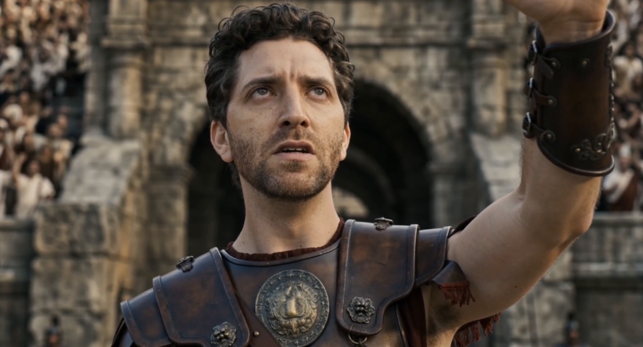 Man with dark curly hair and beard in ancient Roman costume, standing outdoors with stone ruins in background.