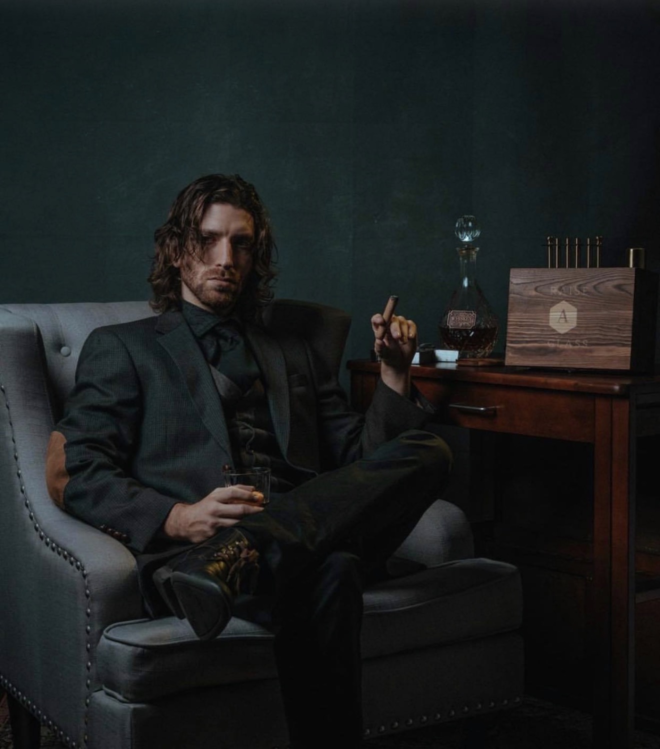 Man with long hair in a dark suit sitting on a sofa, holding a glass, with a wooden cabinet and decorative items behind him.