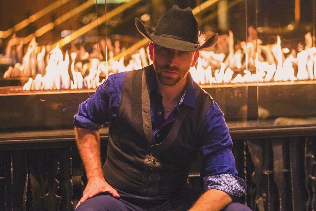 Man wearing a hat and vest sitting near a fire with flames in the background.