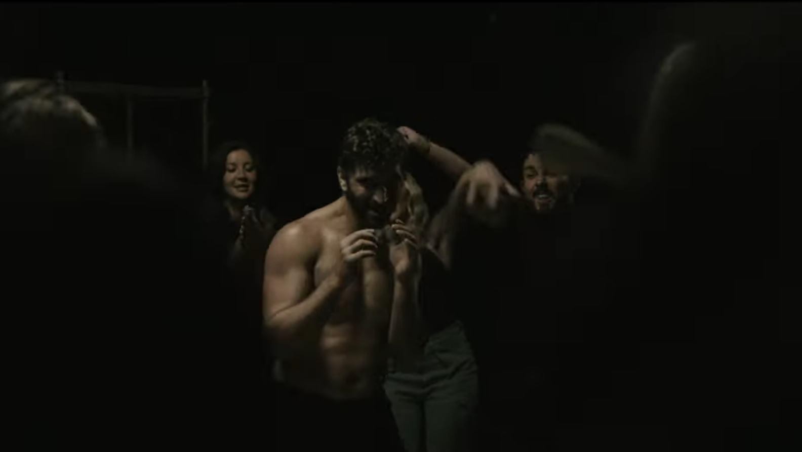 A shirtless man holding a small object, surrounded by smiling people in a dark setting.