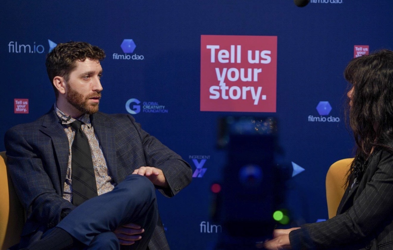 Two people seated in conversation with a blue backdrop and a pink sign that says 'Tell us your story.'