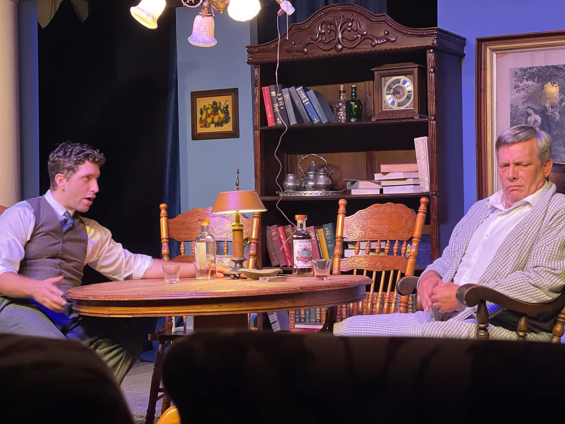 Two men sit at a round table in a room with bookshelves, lamps, and framed pictures, engaged in conversation.