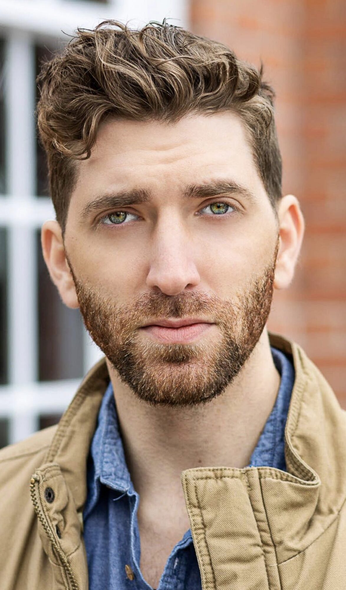 Portrait of a young man with curly hair and a beard, wearing a tan jacket and blue shirt, outdoors.