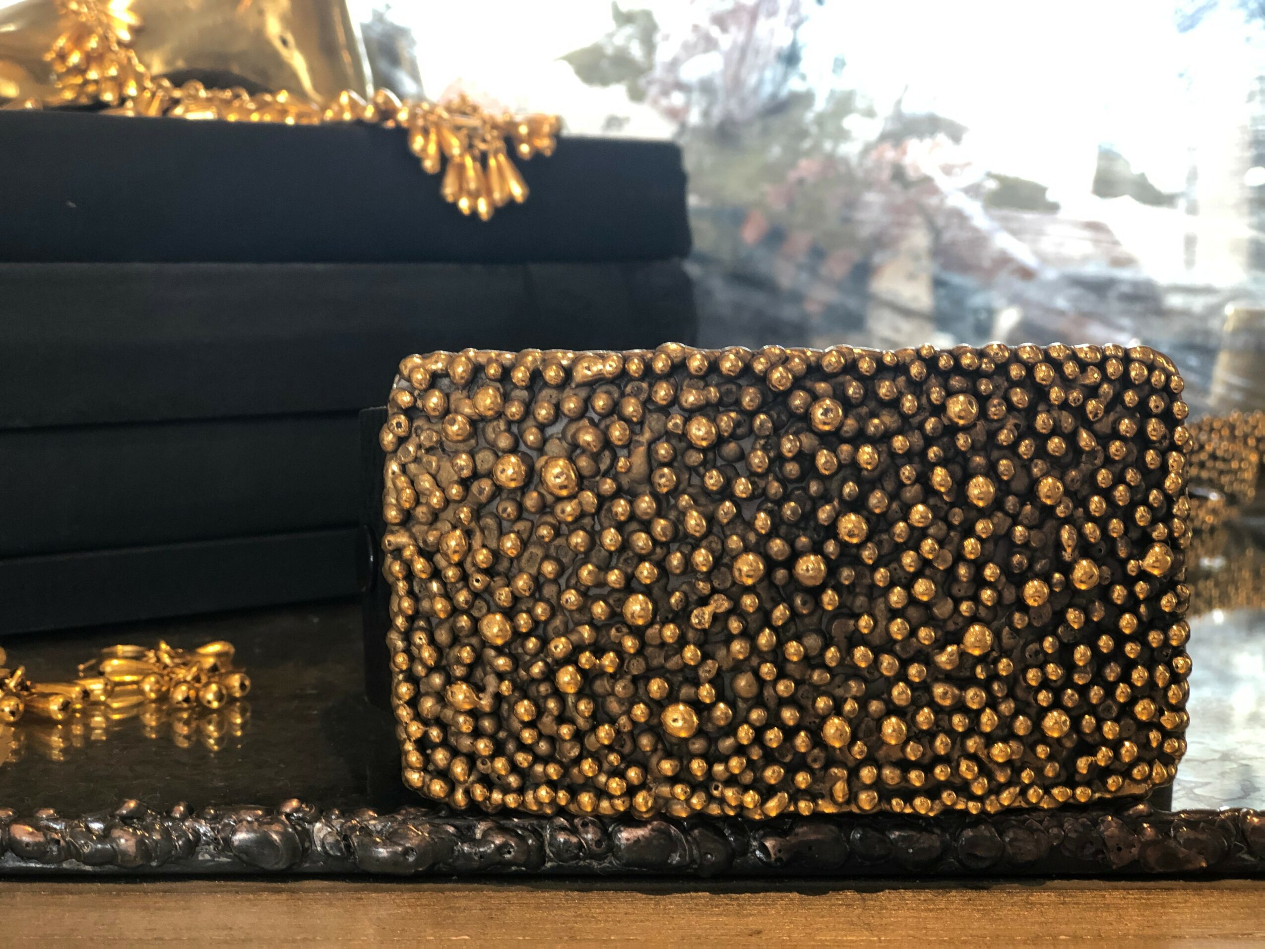 Black clutch purse decorated with gold beads, placed on a dark surface with a black box behind it.