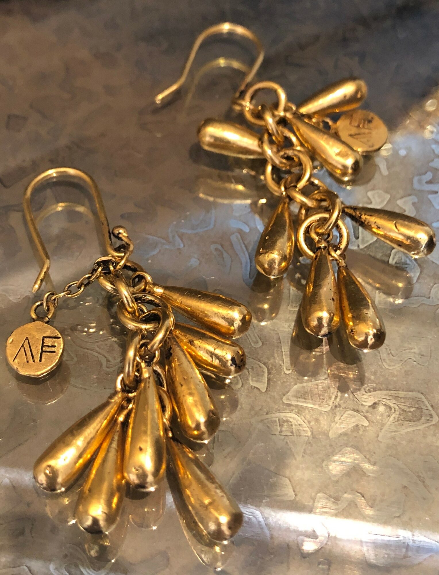 Gold earrings with multiple teardrop-shaped charms hanging from a chain, and a small round charm with initials 'AF'.