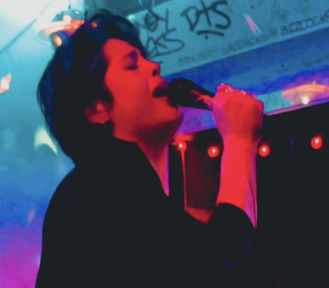 Person singing into a microphone in a colorful, graffiti-covered environment with vibrant lighting.