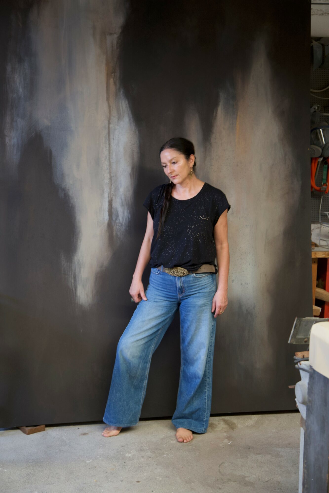 Woman standing against a dark, textured wall, wearing a black top and blue jeans, looking down.