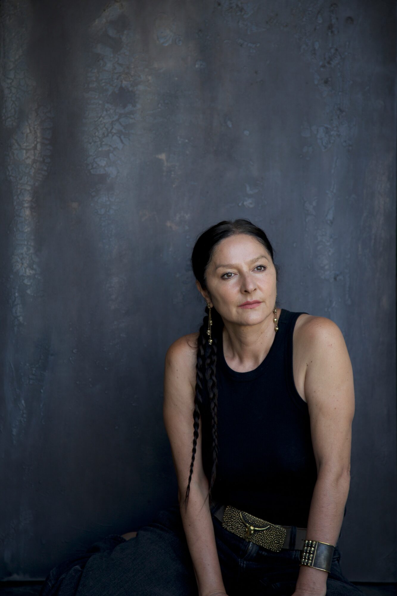 Woman with long braided hair wearing a black sleeveless top and belt, sitting against a dark textured wall.