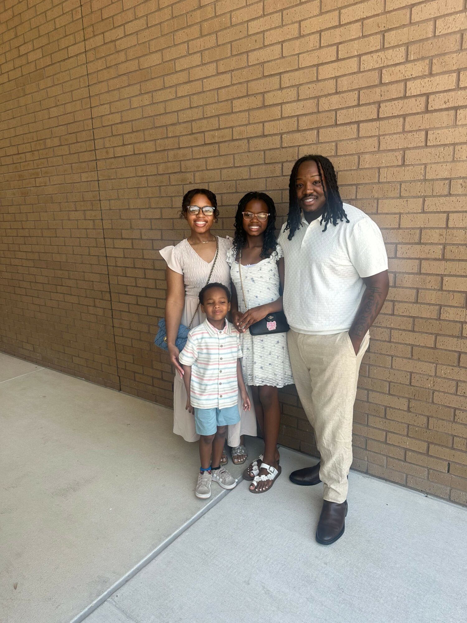 Four people standing against a brick wall, smiling, with a concrete sidewalk in front.