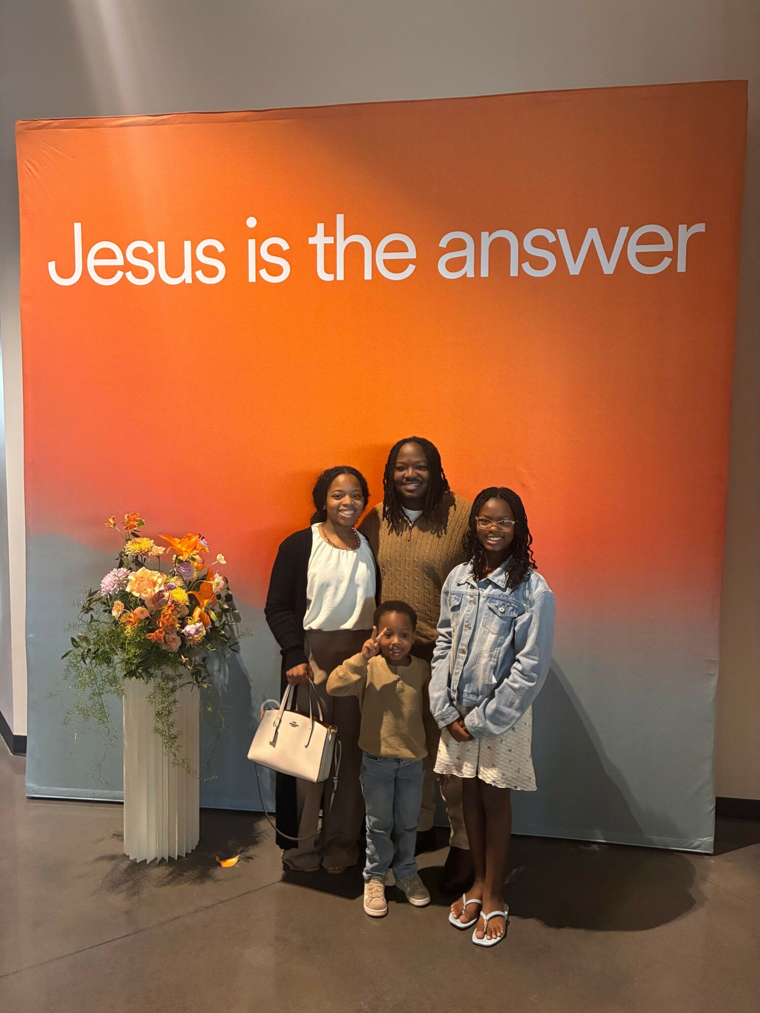 Three women stand in front of a backdrop with orange and blue gradient, with text 'Jesus is the answer'.