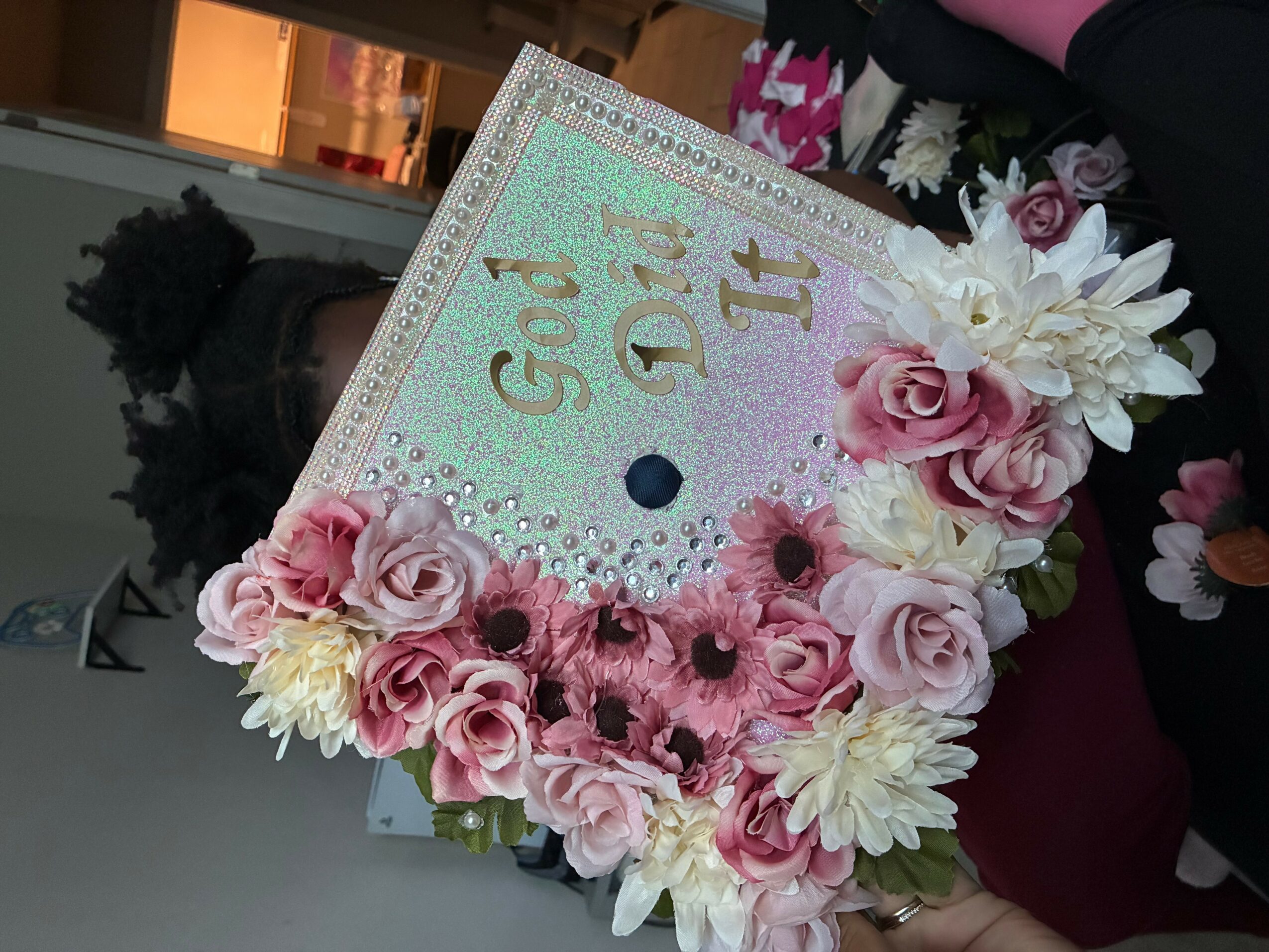 Decorated graduation cap with pink and white flowers, pearls, and the words 'Grad Day' in gold and green, on a table.