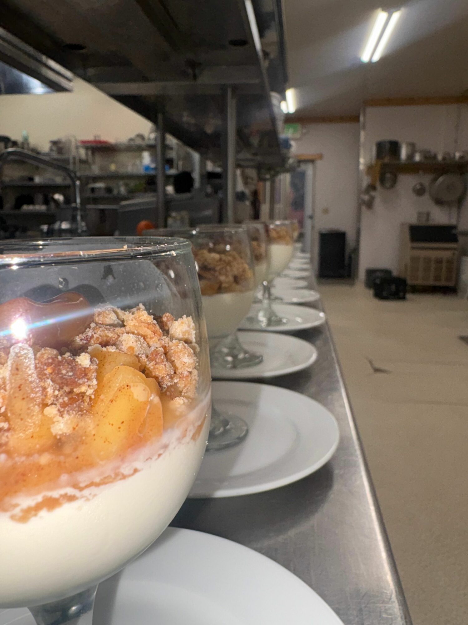 A row of dessert glasses with layered desserts on a counter, with plates and a kitchen background.