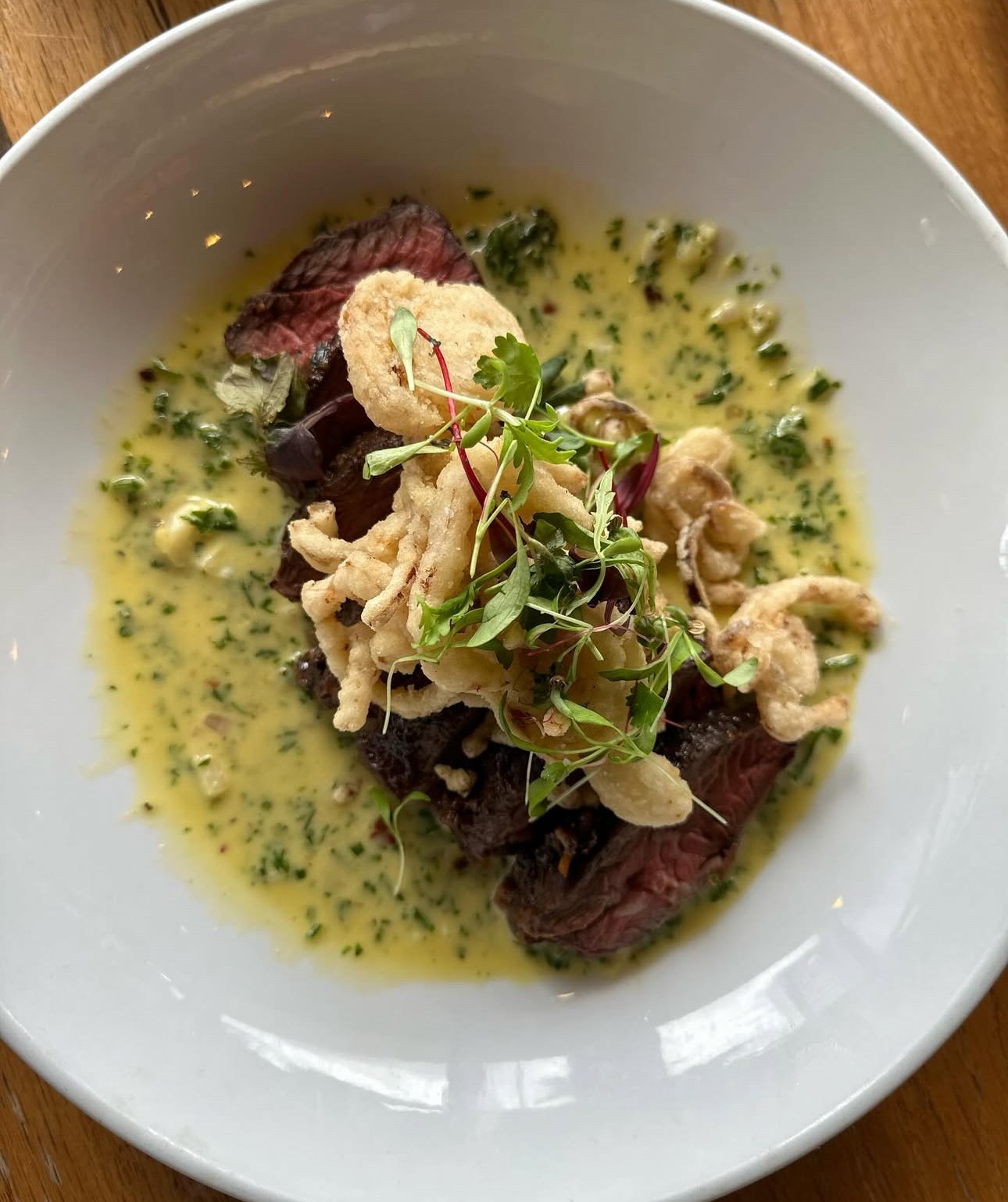 Sliced cooked beef topped with microgreens and crispy elements, served with a green herb sauce in a white bowl.