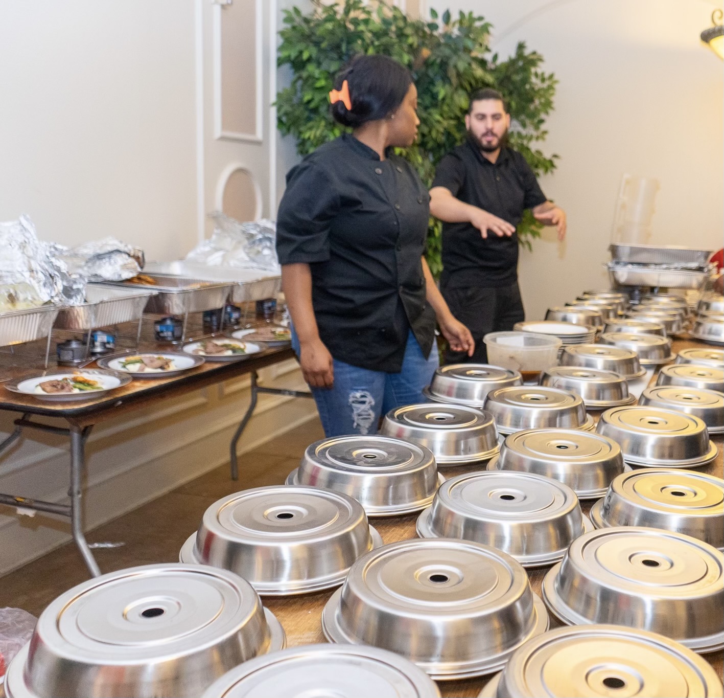 Two people preparing food in a room with tables of covered dishes and food containers, with a large plant in the background.