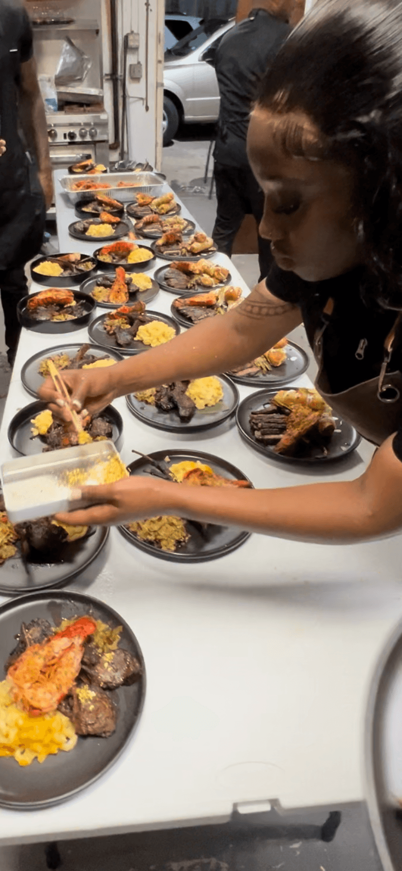 Girl serving food at a table with plates of rice, meat, and vegetables outdoors, cars visible in the background.