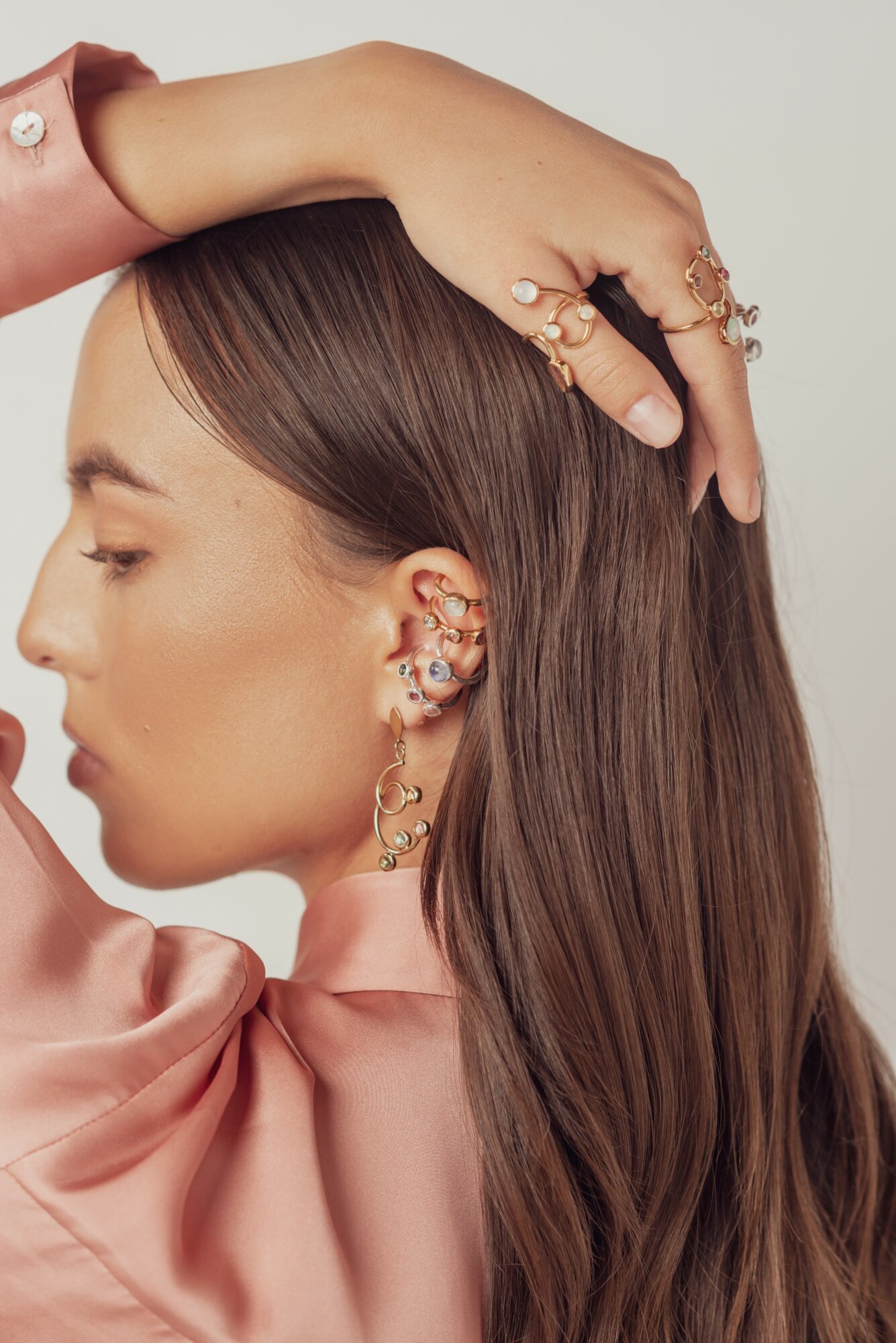 Side profile of a woman with long brown hair, wearing multiple earrings and rings, dressed in pink satin fabric.