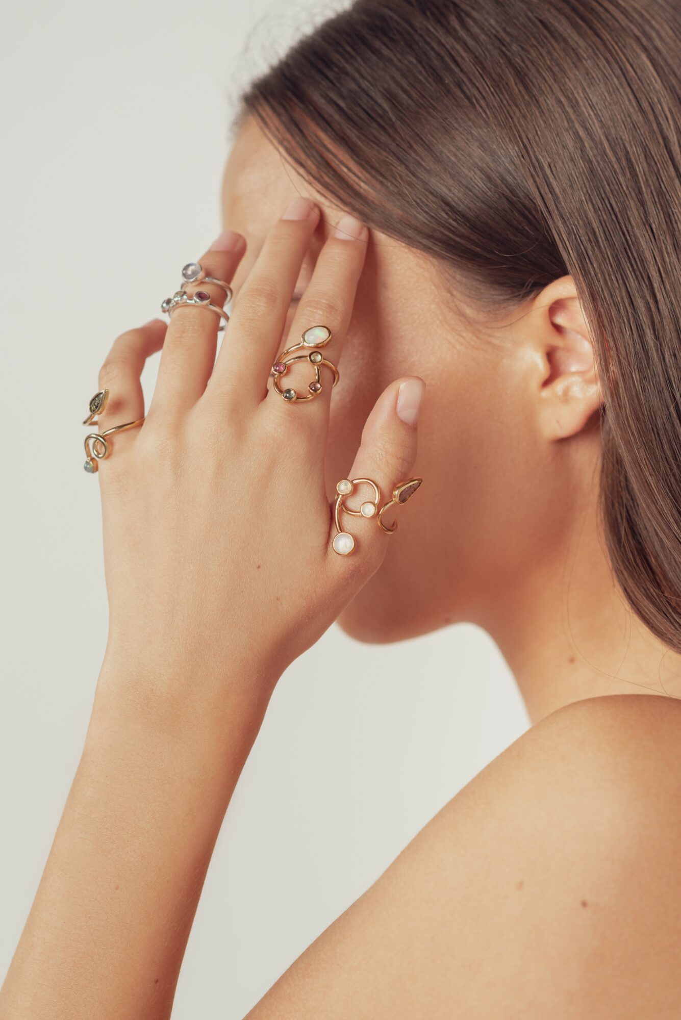 Person with multiple rings on fingers, covering face with hand, against a plain background.