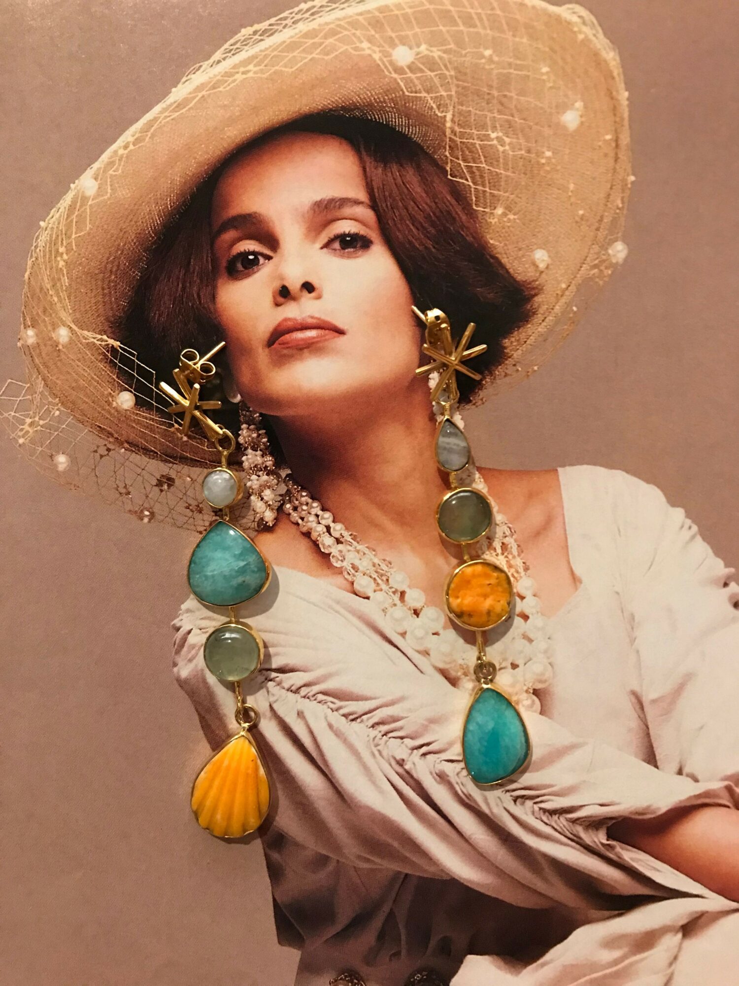 Woman wearing a large straw hat with decorative elements, lying on her side, with a colorful beaded necklace, against a plain background.